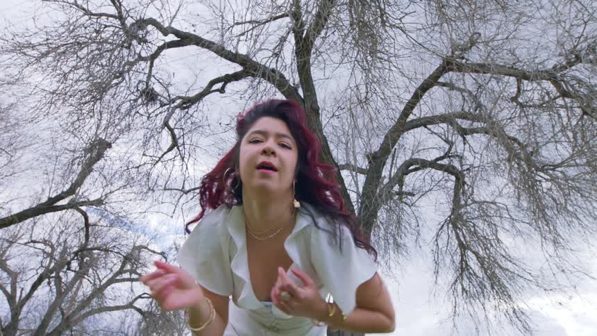 A woman with red hair and a white dress is jumping with her arms up and behind her is a leafless tree