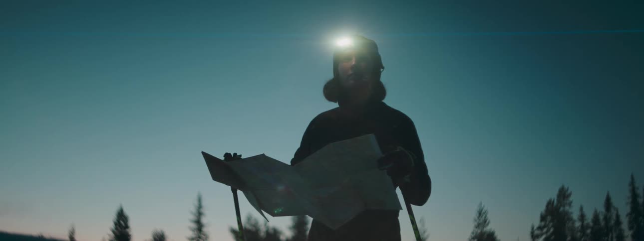 A person is holding a map and opens it and there is a flashlight on his head and trees in the background