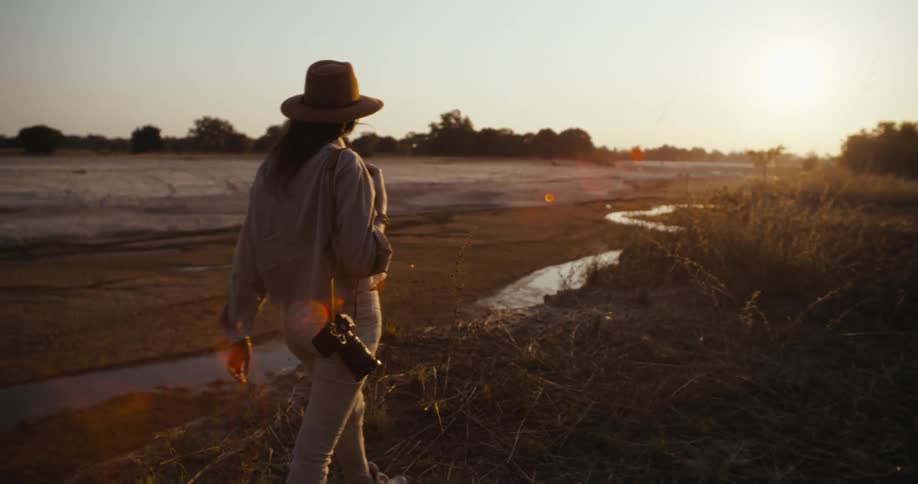 A woman in a hat carrying a camera watches the sun set over a dry field