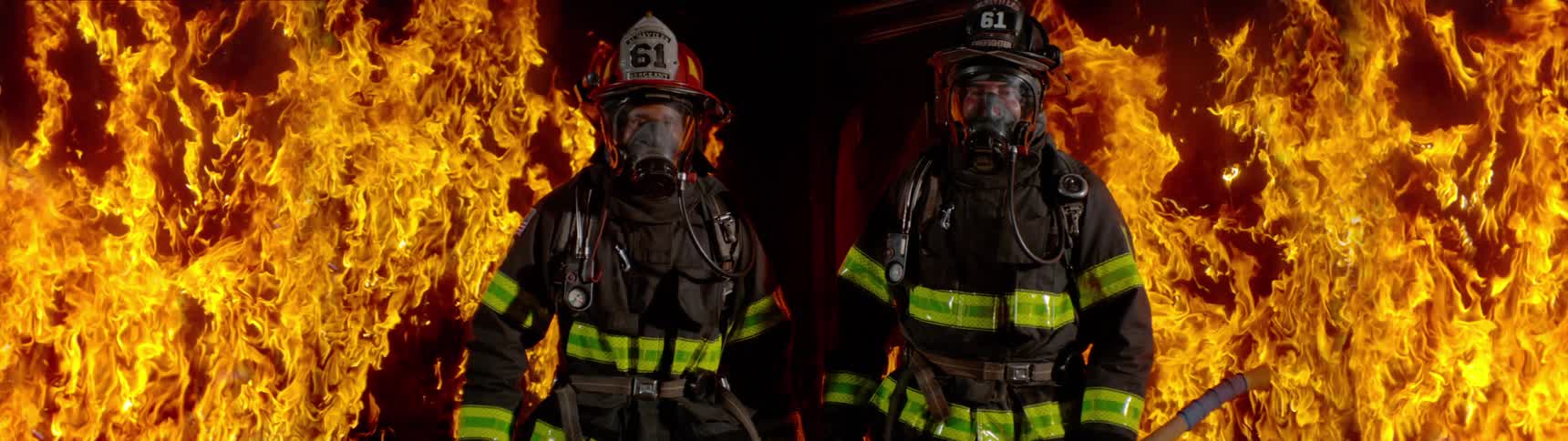 Two firemen stand near a raging fire at night