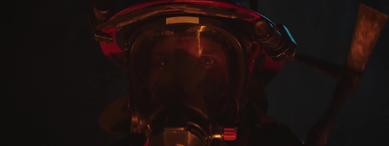 A firefighter in a helmet and mask is lit by a fire in a dark setting