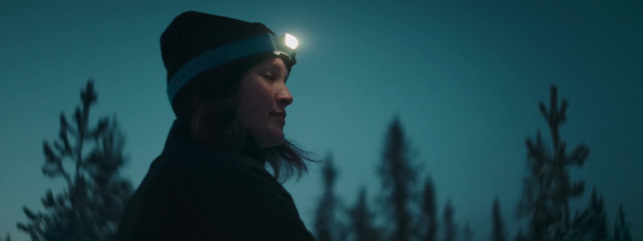 Woman wearing a headlamp walks on skis in a snowy forest
