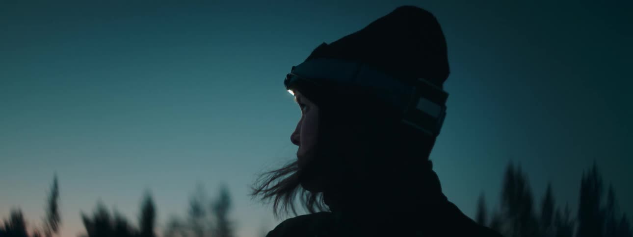A woman with a headlamp looks around in the forest at nightfall