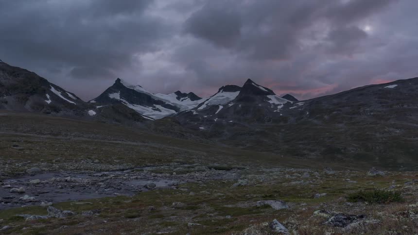 A valley with clouds rolling over jagged mountains in the background time lapsed from morning to evening