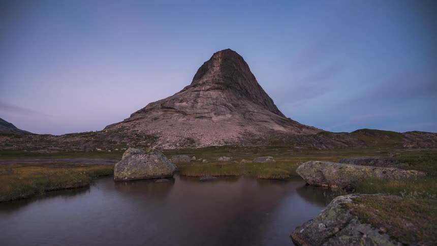 Thin clouds in a blue sky quickly moving over a mountain peak by a lake time lapse day to evening