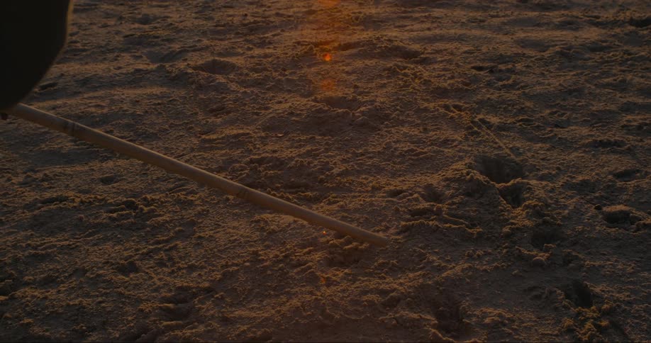 A person holding showing animals tracks a stick on the dirt floor during sunset