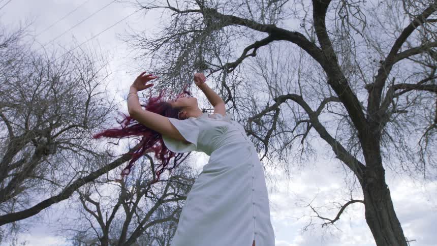 Woman wearing a white dress shakes her colorful hair in front of barren trees