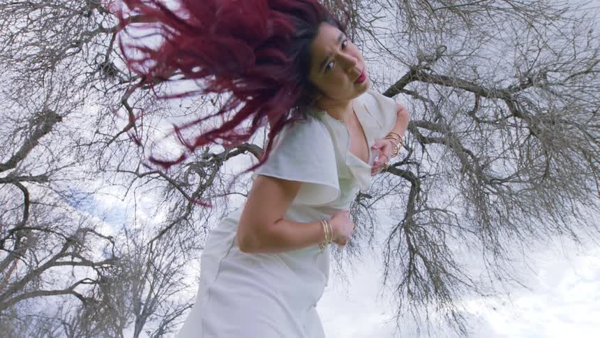 Woman in a white dress shakes her long colorful hair in front of a barren tree