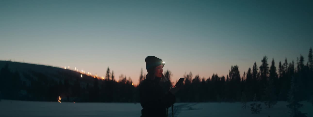 A woman with a flashlight on her forehead in a snowy field checking her smart phone