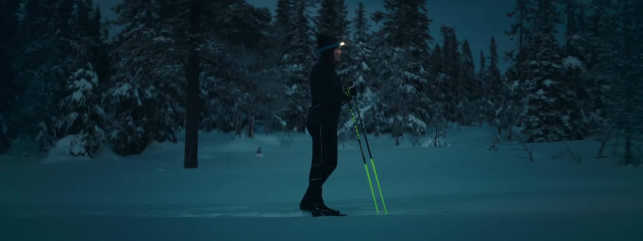 A skier with a headlamp stands in a field of snow surrounded by trees