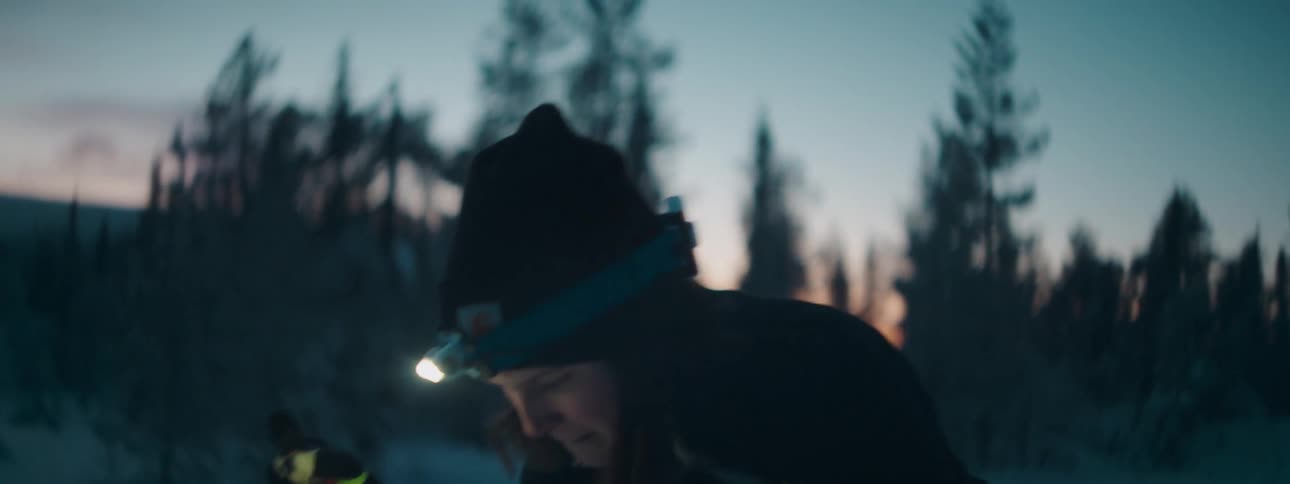 Lady walking on snow with skis and headlamp at sunset