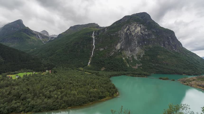Waterfalls and steep mountains are surrounded by lush foliage and a turquoise lake on a cloudy day