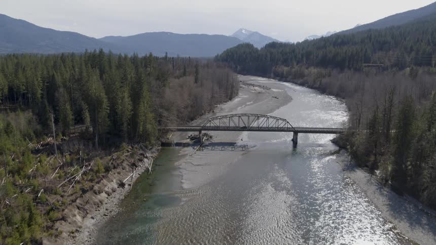 A SUV crosses a bridge over a rushing river surrounded by woods and mountains in the distance