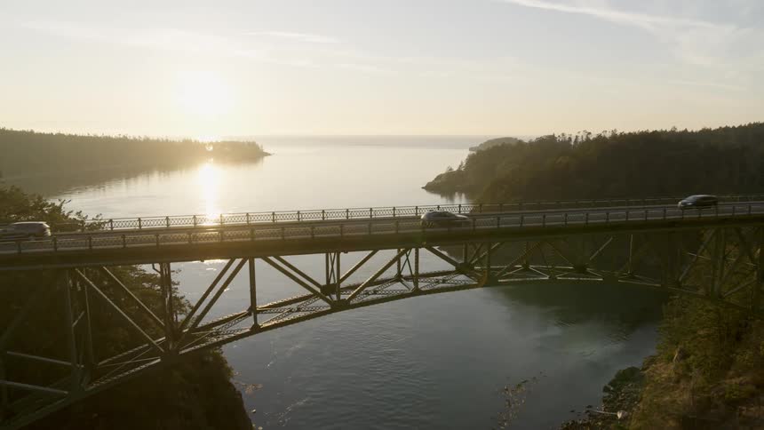 Reveal of a backlit bridge span high over water as cars and SUVs drive across