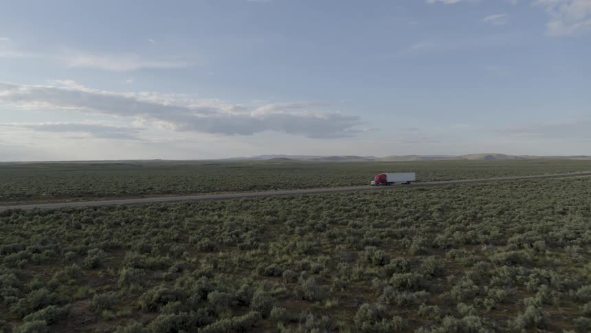 A semi truck travels cross country along a rural highway surrounded by sagebrush bathed in soft light