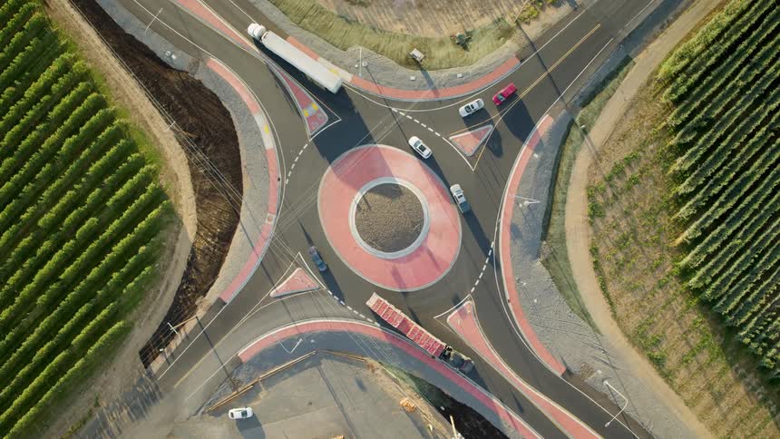 Semi trucks drive in opposite directions through a roundabout between fruit orchards