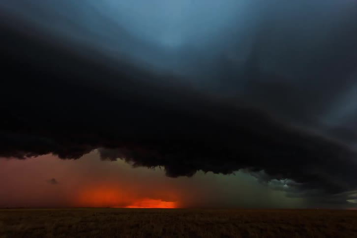 Dark storm clouds and a supercell storm moves over the land as lightning flashes and strikes the ground