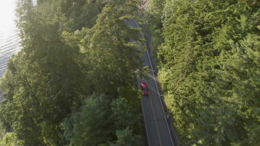 A red car drives on a beautiful cliffside highway bathed in warm sunshine