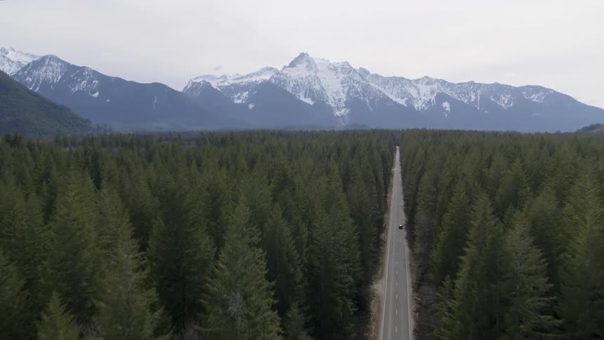 Orbiting reveal of a car driving on a country road through dense forest with snow capped mountains in the distance