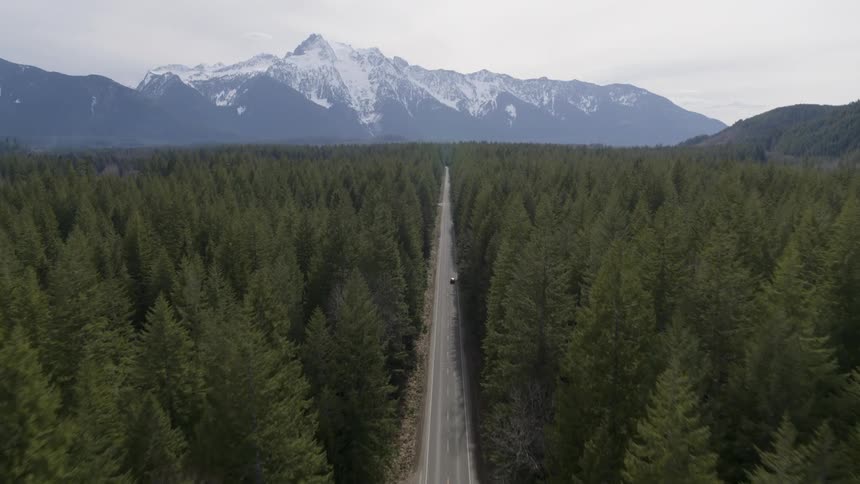 Following a car driving towards a dramatic mountain range on a highway through dense forest