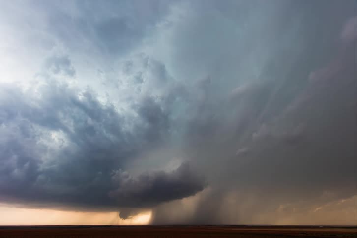 A supercell storm pours rain and moves across the land as lightning flashes