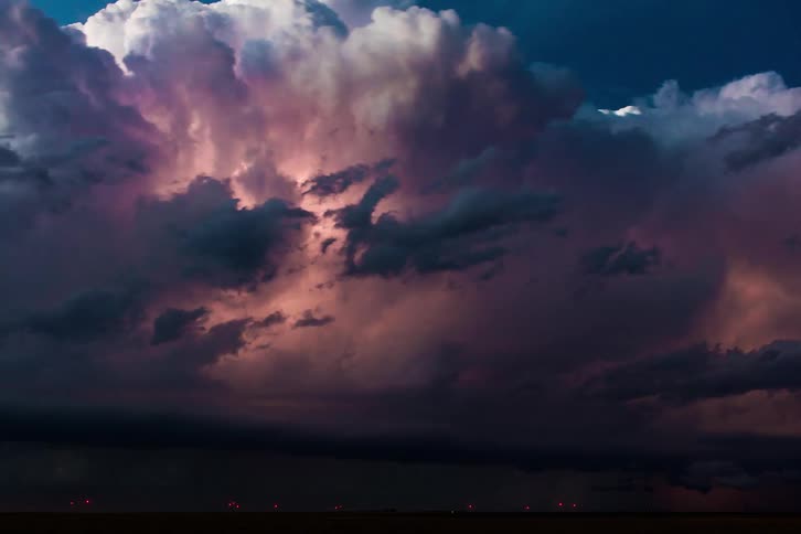 Cumulus clouds float above dark storm clouds pouring rain on the ground as lightning strikes and flashes as they fill the dark sky