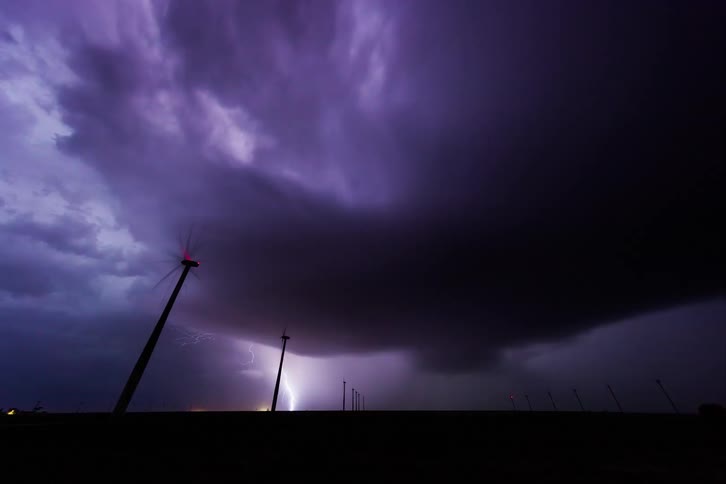 A windmill farm and on top of a huge storm with heavy clouds and several lightning strikes falling to the ground