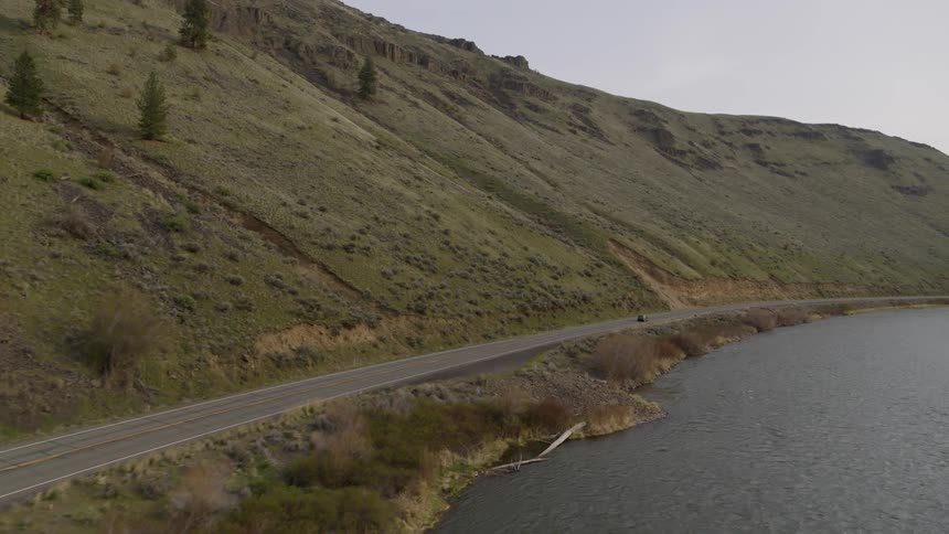 A small SUV driving on an empty highway between a rugged hillside and flowing river