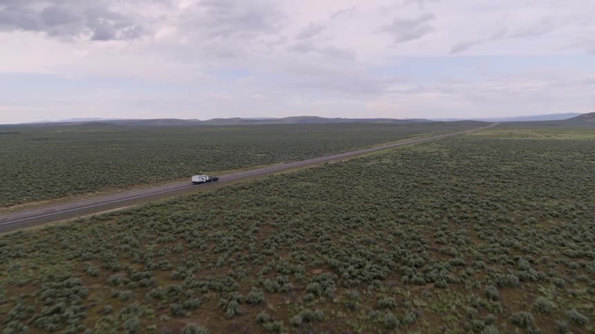 A pickup tows a camper along open highway towards foothills in the distance
