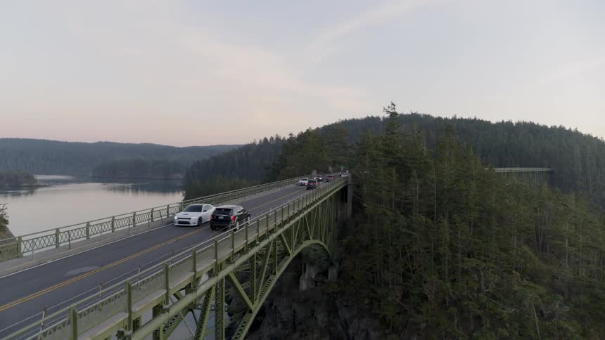 Evening commuters drive along a narrow bridge over reflective water far below