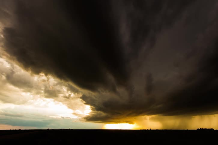 Dark storm clouds move across the sky as rain pours onto the ground and the sun glows on the horizon