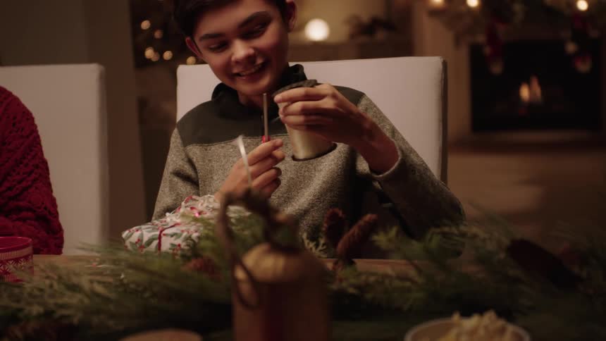 Portrait of two children with one of them taking scissors and cutting a gift ribbon while sitting at a dining table at night
