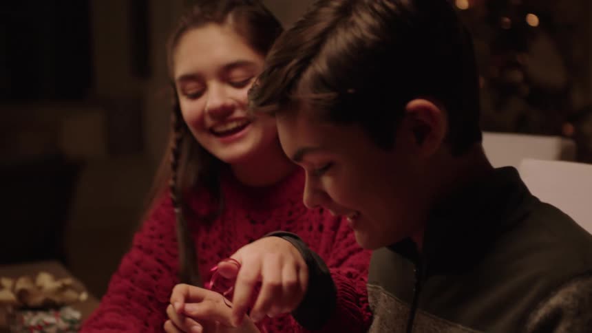Two teenagers a boy and a girl laugh as they wrap a christmas gift together