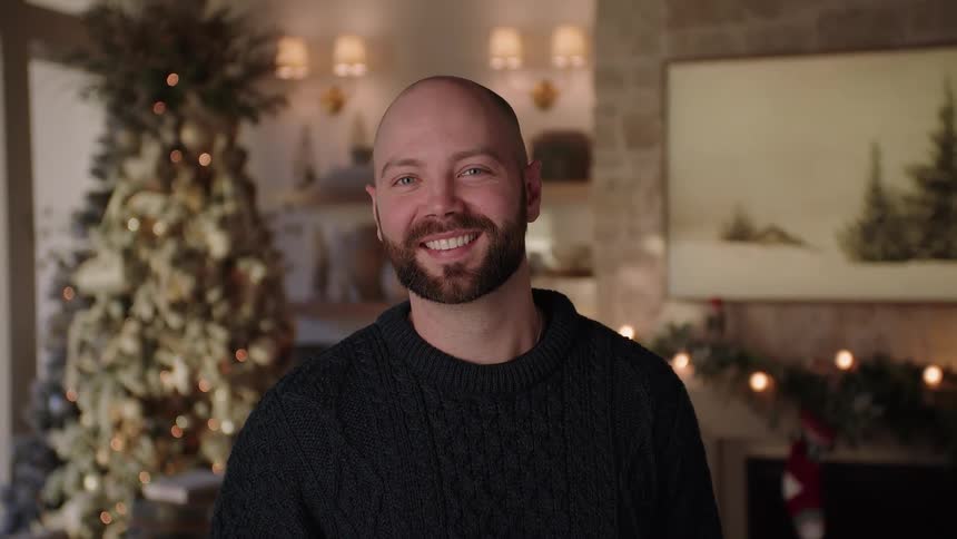 Man with a black sweater and a beard smiles at camera in front of christmas tree