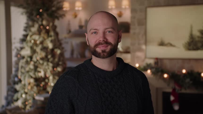 Man with a beard and a black sweater stands in front of the christmas tree smiling