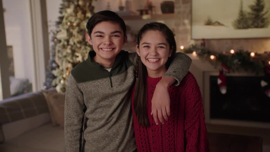 Brother throws his arm around his sister as they stand smiling in front of christmas tree