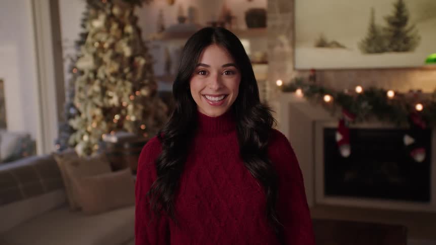 Woman with dark wavy hair and a red sweater smiles at camera in the living room in front of christmas tree