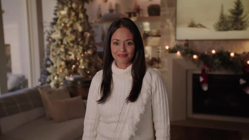 A woman in a white sweater poses in a living room in front of a decorated christmas tree