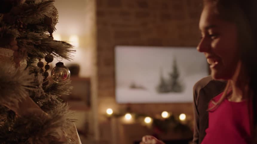 A teenage boy and girl are smiling as they put ornaments on a christmas tree with some lights