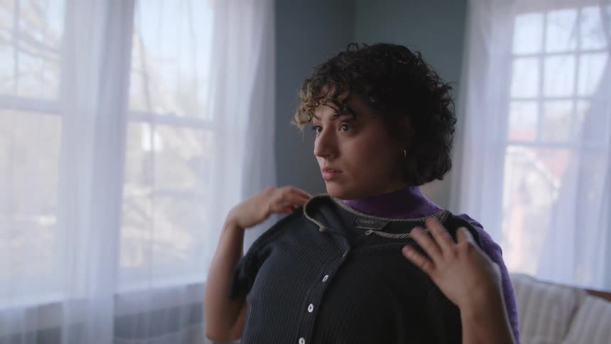 A woman taking clothes from a bed and placing them on her torso and making expressions