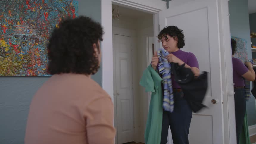 A woman shows her husband how several clothes items look on her asking him for advice on what to wear