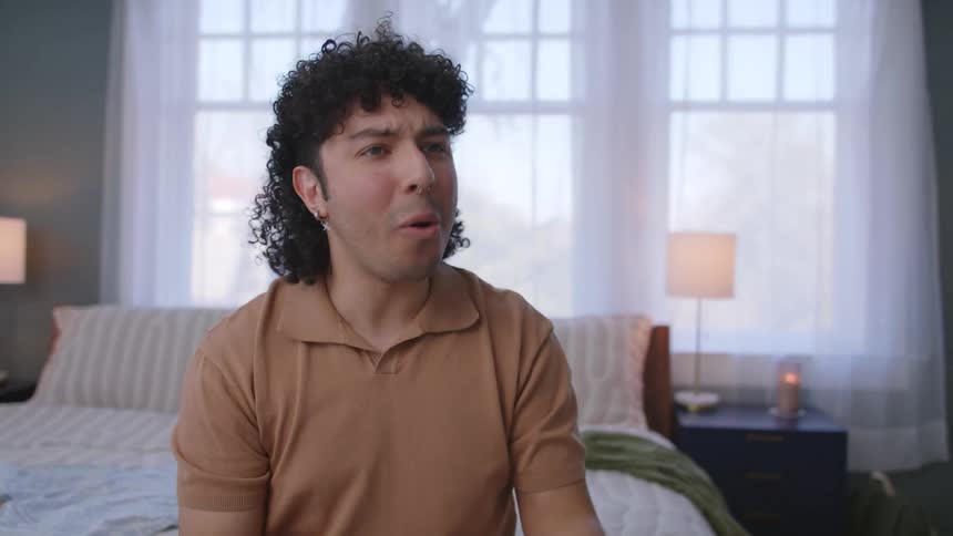 A man is talking and smiling with someone in front of him while sitting on a bed
