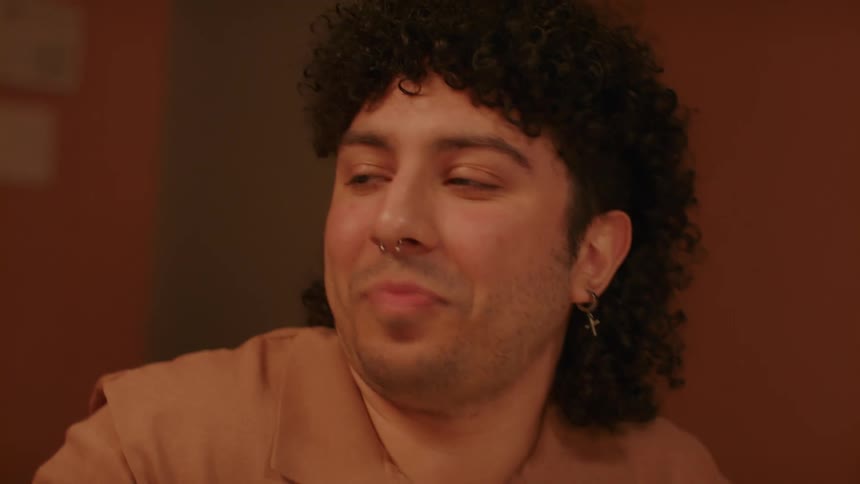 A man with curly hair a nose piercing earrings and a brown polo shirt sits at a table smiling