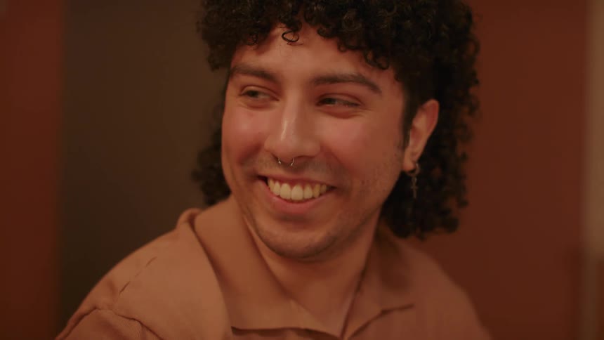 Man with curly hair earrings and a nose piercing sits at table talking to people