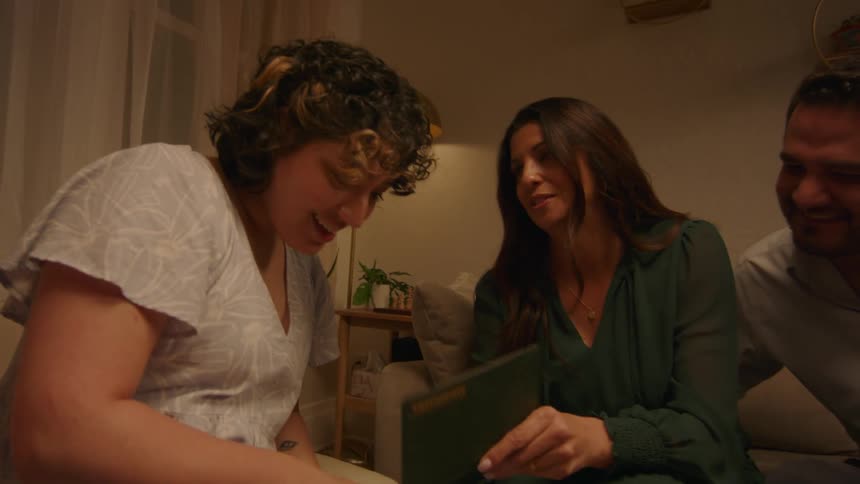 Three people sitting in a living room talk and laugh as they show each other photos from a book