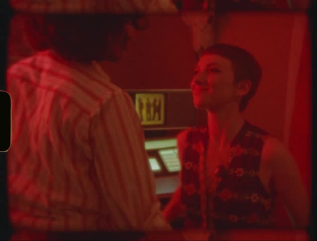 A woman pulls a seated man by the hand and they both begin to dance in a room with red lights