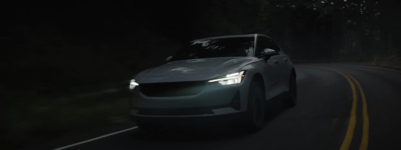 Sleek gray car with white headlight driving on a dimly lit highway through the woods
