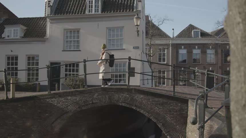 A person walks on a bridge over a river in a town with old buildings and a clear sky