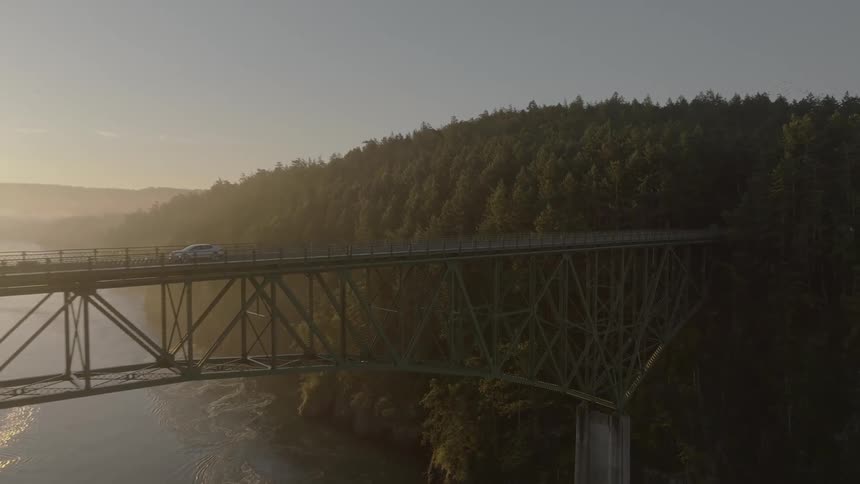 Sun shines from near horizon as car drives across bridge over river bordering forest covered mountain