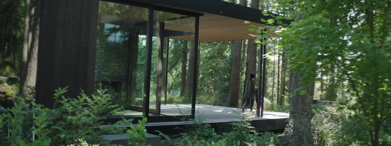 A woman and a young daughter walk out of a glass house and into the woods hand in hand